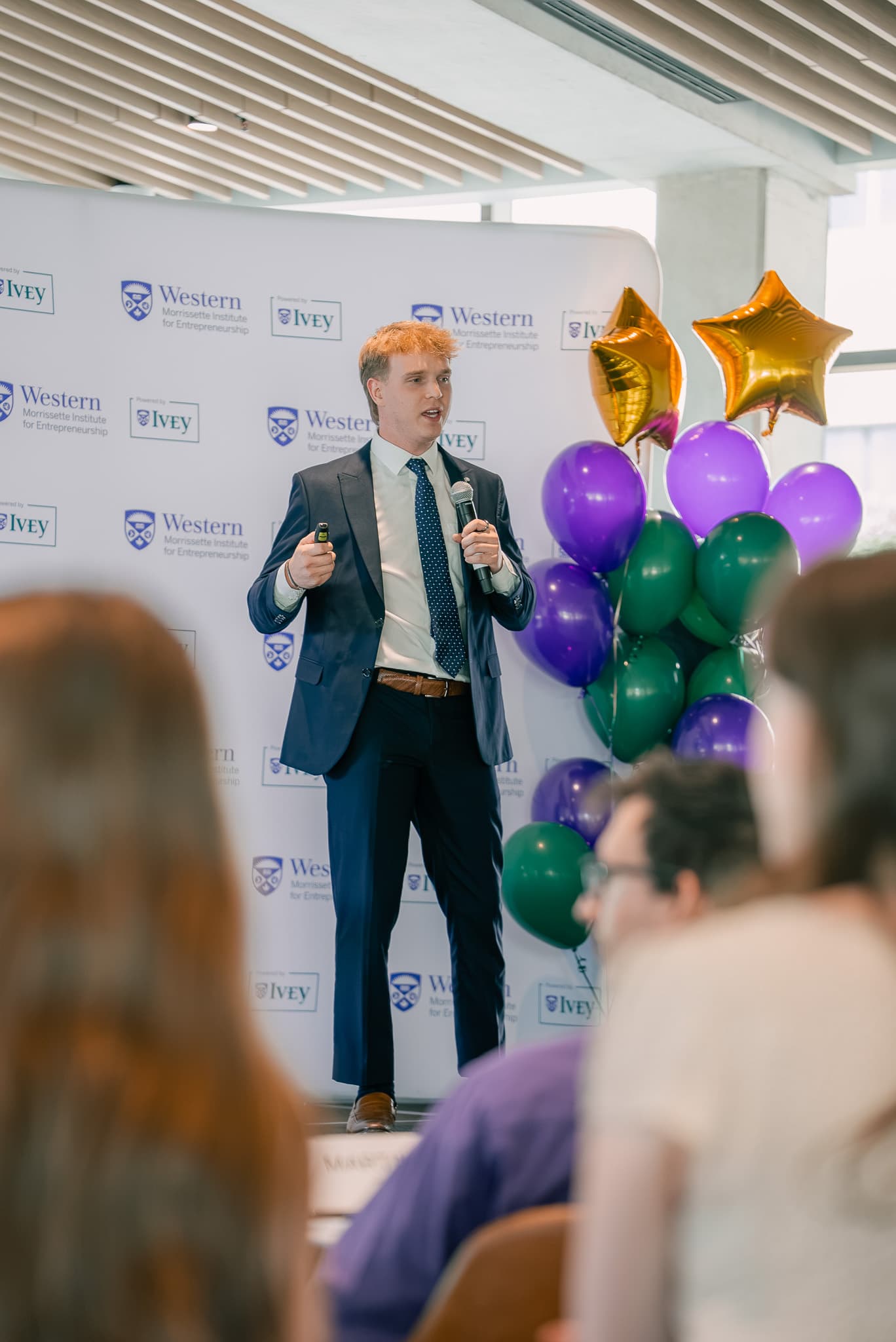 A presenter speaking into a microphone beside purple, green, and gold balloons.